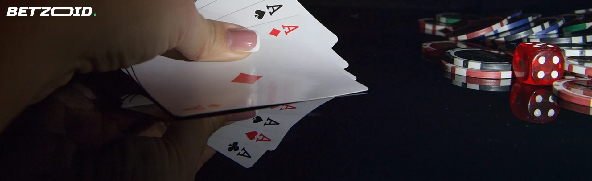 A hand fanning out playing cards with poker chips and dice in the background on a dark table.